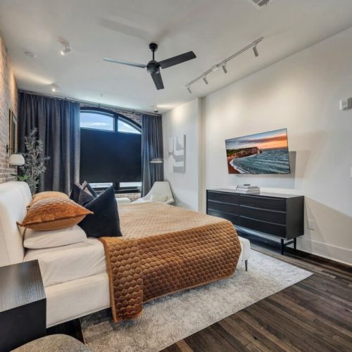 Cozy and stylish bedroom featuring a comfortable bed, soft layered bedding, a smart TV, and a large window with blackout curtains for a relaxing stay.