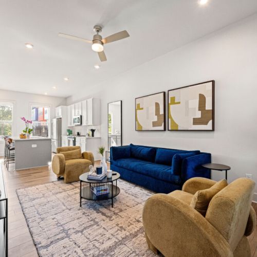 Wide view of the main living space showcasing the plush navy sofa, two cozy accent chairs, modern artwork, and an open layout connecting the living room and kitchen for easy entertaining.