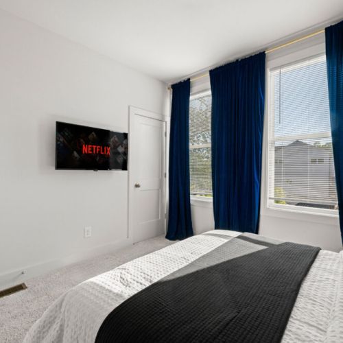 Bedroom 2 - Bright room featuring a comfortable bed, a Smart TV, and large windows with blinds and navy curtains. The spacious layout offers a relaxing retreat with plenty of natural light.