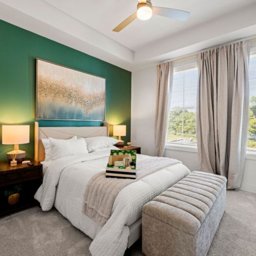 Bedroom 1 - Spacious queen bedroom featuring a plush queen-size bed, ceiling fan, bedside lamps, and large windows with blinds and curtains. An upholstered bench adds convenient seating at the foot of the bed.